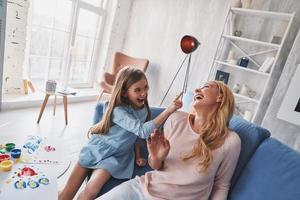 vista superior de una linda niñita tratando de darle a su joven madre una cara pintada y riendo mientras pasa tiempo sin preocupaciones en casa foto