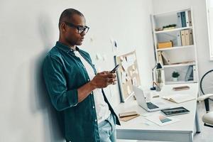 Business is his life. Handsome young African man in shirt using smart phone while standing in the office photo