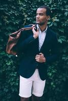 Confident in his perfect style. Handsome young African man in smart casual wear carrying bag on shoulder and looking away while standing against green plant background outdoors photo