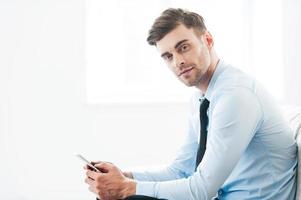 Always staying connected. Side view of confident young businessman holding mobile phone and looking at camera while sitting on sofa photo