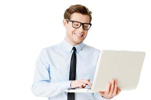 es profesional. un joven apuesto con camisa y corbata trabajando en una laptop y sonriendo mientras está aislado en blanco foto