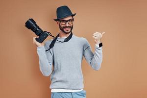 joven feliz con ropa informal mirando la cámara y señalando el espacio de copia mientras se enfrenta a un fondo marrón foto