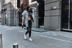 Great look. Full length of beautiful young woman in jeans jacket keeping hand in hair while walking down the street photo