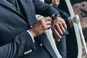 Always in time. Close up of young man in full suit checking the time while standing in front of the mirror indoors photo