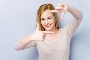 Gesturing finger frame. Beautiful young woman looking at camera and gesturing finger frame while standing against grey background photo