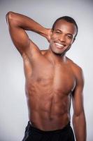 Feeling strong and confident. Young African man holding hand behind head looking at camera and keeping arms crossed while standing isolated on grey background photo