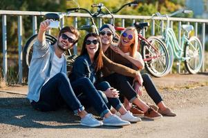 capturando diversión. grupo de jóvenes sonrientes que se unen entre sí y se hacen selfie por teléfono inteligente mientras se sientan al aire libre junto con bicicletas en el fondo foto