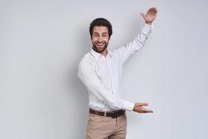 Happy young man in white shirt looking at camera and gesturing while standing against gray background photo