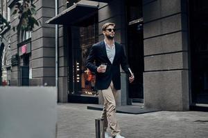 Handsome businessman. Handsome young man in smart casual wear carrying disposable cup while walking through the city street photo