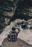 His best shots. Top view of young modern man photographing while sitting on the rock with the river below photo