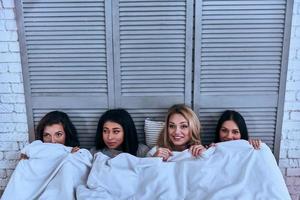 hora de dormir. vista superior de cuatro hermosas mujeres jóvenes que cubren con una manta blanca y sonríen mientras están acostadas en la cama foto