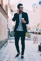 hablando con un amigo toda la longitud de un joven apuesto con ropa informal elegante que lleva una taza de café y habla por teléfono móvil mientras camina por la calle foto