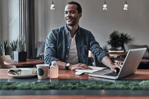 Best place to work. Handsome young man using computer and smiling while sitting in the cafe photo