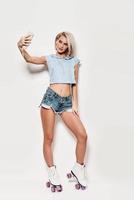 buena autofoto Longitud total de mujer joven en ropa casual tomando selfie y sonriendo mientras patina sobre fondo gris foto