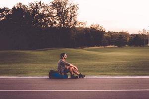 Tired traveler. Side view of handsome young man sitting on his backpack at the side of the road and looking thoughtful photo