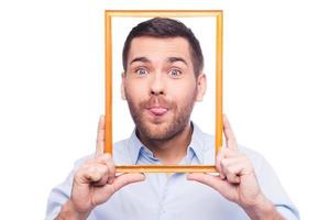 Playful portrait Handsome young man in shirt sticking his tongue out and holding picture frame in front of his face while standing against white background photo