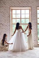 Making sure she looks perfect. Full length of bridesmaids helping bride with her wedding dress while standing near the window together photo