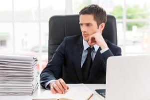 Missing the deadlines. Thoughtful young man in shirt and tie looking at the stack of paperwork and holding head on chin while sitting at his working place photo