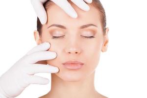 Preparation before facial surgery. Beautiful young woman keeping eyes closed while doctor in medical gloves examining her face isolated on white photo