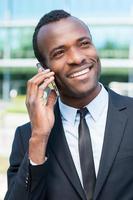 hablando de negocios. alegre joven africano con ropa formal hablando por teléfono móvil y sonriendo mientras está de pie al aire libre foto