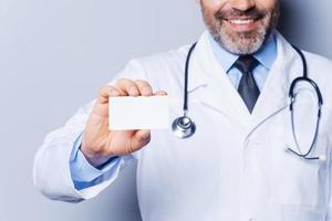 Doctor holding copy space. Close-up of confident mature doctor showing his business card and smiling while standing against grey background photo