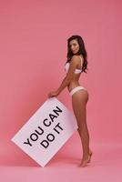 Full length of attractive young woman holding a poster and smiling while standing on tiptoe against pink background photo