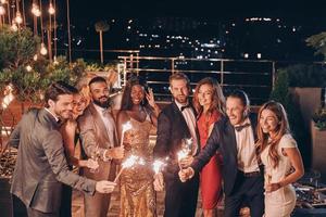 grupo de personas hermosas en ropa formal sosteniendo bengalas y sonriendo mientras pasan tiempo en una fiesta al aire libre foto