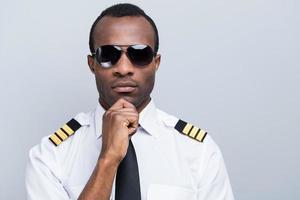 Confident and experienced pilot. Confident African pilot in uniform holding hand on chin while standing against grey background photo