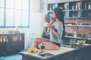 mañana sin preocupaciones. bella joven mestiza sosteniendo una taza de café y hablando por teléfono móvil con una sonrisa mientras se sienta en el escritorio de la cocina foto