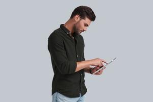 Putting ideas into something real. Serious young man working on his digital tablet while standing against grey background photo