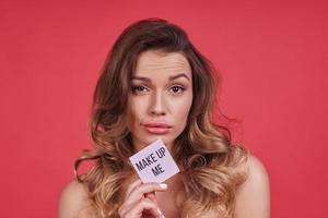 Need some make-up.  Beautiful young woman looking at camera and holding a little poster while standing against pink background photo