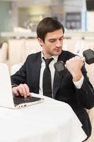 Healthy lifestyle. Confident young man in formalwear sitting at laptop and holding a dumbbell photo