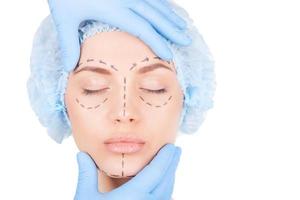 Preparation for facial surgery. Attractive young woman in medical headwear and sketches on face keeping eyes closed while doctor examining her face isolated on white photo