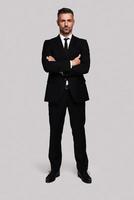 Young and successful. Full length of handsome young man in full suit keeping arms crossed and looking at camera while standing against grey background photo