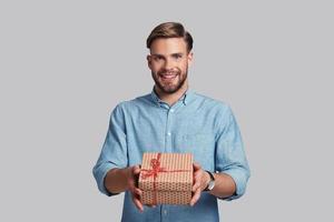 This is for you.  Handsome young man giving gift box and smiling while standing against grey background photo