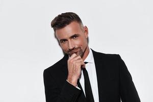 Confident business expert. Handsome young man in full suit keeping hand on chin and looking at camera with smile while standing against white background photo