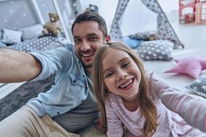 Nice selfie Self portrait of young father and his little daughter smiling while sitting on the floor in bedroom with the tent in the background photo