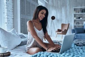 Feeling happy. Attractive young smiling woman using laptop and looking at it while sitting on the bed at home photo