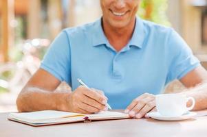 Making some important notes. Close-up of confident mature man writing something in his note pad and smiling while sitting at the table outdoors with house in the background photo