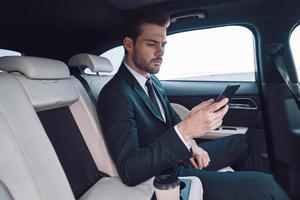 Young perfectionist. Handsome young man in full suit using his smart phone while sitting in the car photo