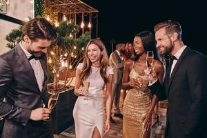 grupo de personas hermosas en ropa formal comunicándose y sonriendo mientras pasan tiempo en una fiesta de lujo foto