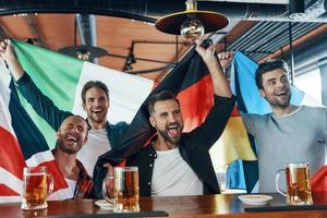 Happy young men covered in international flags drinking beer and watching sport game while sitting in the pub photo