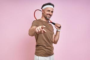 Happy mature man carrying tennis racket and pointing camera while standing against pink background photo