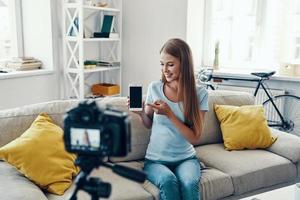 hermosa mujer joven sonriendo y señalando espacio de copia en su teléfono inteligente mientras hace videos de medios sociales en casa foto