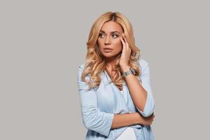 Something bothers her. Thoughtful young woman looking away and touching her face with hand while standing against grey background photo