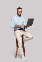 May I help Full length of good looking young man smiling and using computer while sitting on stool against grey background photo