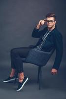 Take a closer look at my style. Confident young handsome man adjusting his glasses and looking at camera while sitting in chair against grey background photo