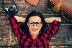 Ready to a big trip. Top view of beautiful young woman in eyewear lying on the hardwood floor and smiling while suitcase and camera laying near her photo