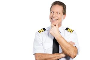 apasionado por el cielo. piloto masculino pensativo en uniforme sosteniendo la mano en la barbilla y mirando hacia arriba con una sonrisa mientras está de pie contra el fondo blanco foto