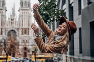 Great look. Attractive young smiling woman in hat and coat taking selfie while spending time in the city photo